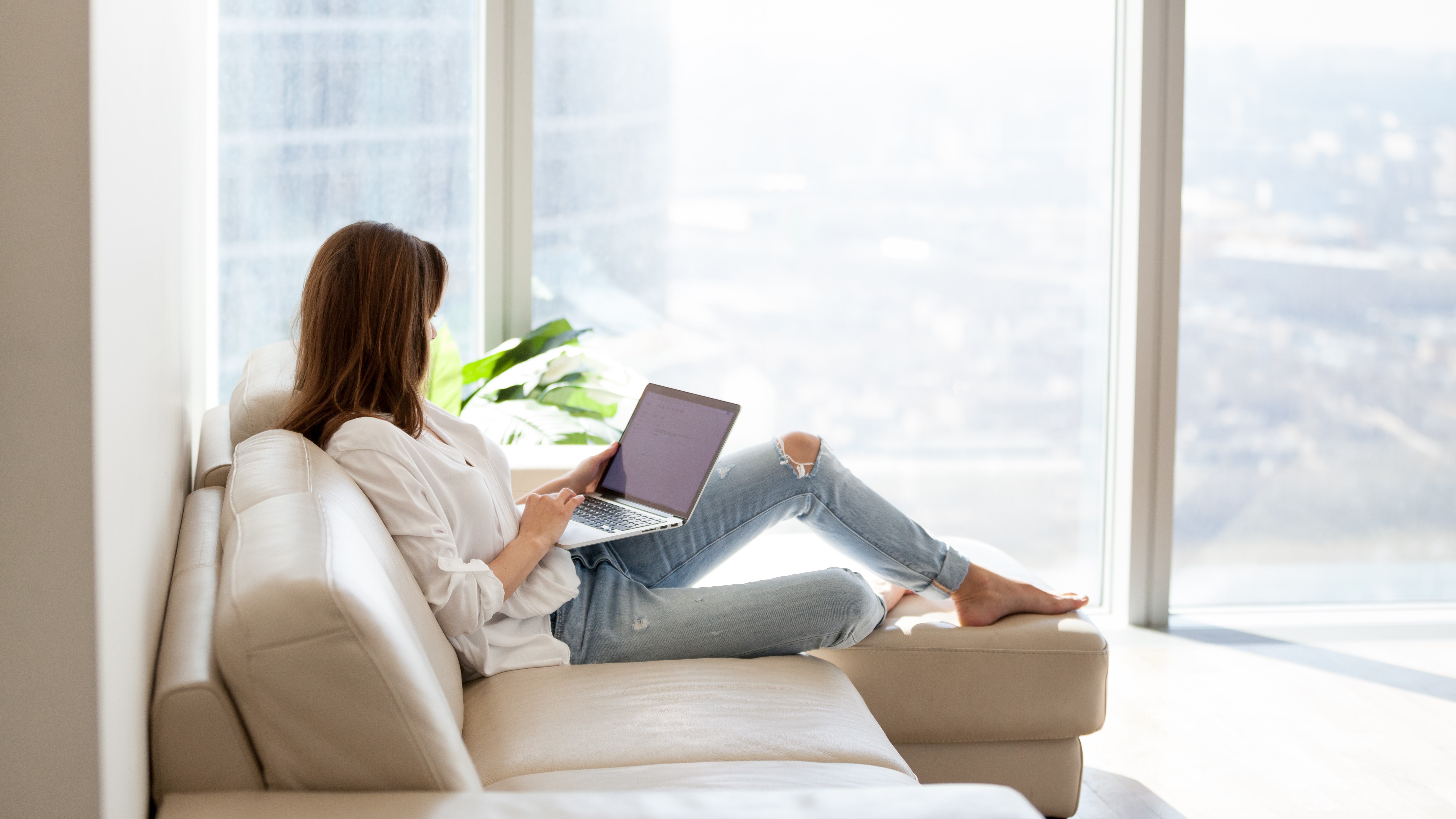 Женщина отдыхает на диван, держит ноутбук на коленях и пользуется им