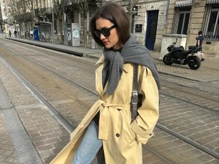 French fashion influencer Anne-Laure Mais in Paris wearing blue jeans, a classic trench, sunglasses, a gray knit tied at her shoulders, and black boots.