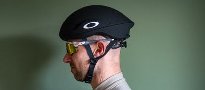 A rider wearing a black Oakley Velo Mac helmet in front of a green wall 