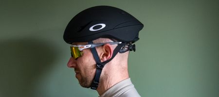 A rider wearing a black Oakley Velo Mac helmet in front of a green wall 