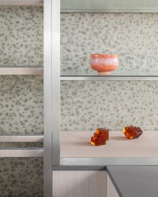 Close up view of a metal shelving unit against a patterned wall at the Turner Contemporary shop. There is an orange bowl and two amber coloured pieces on display