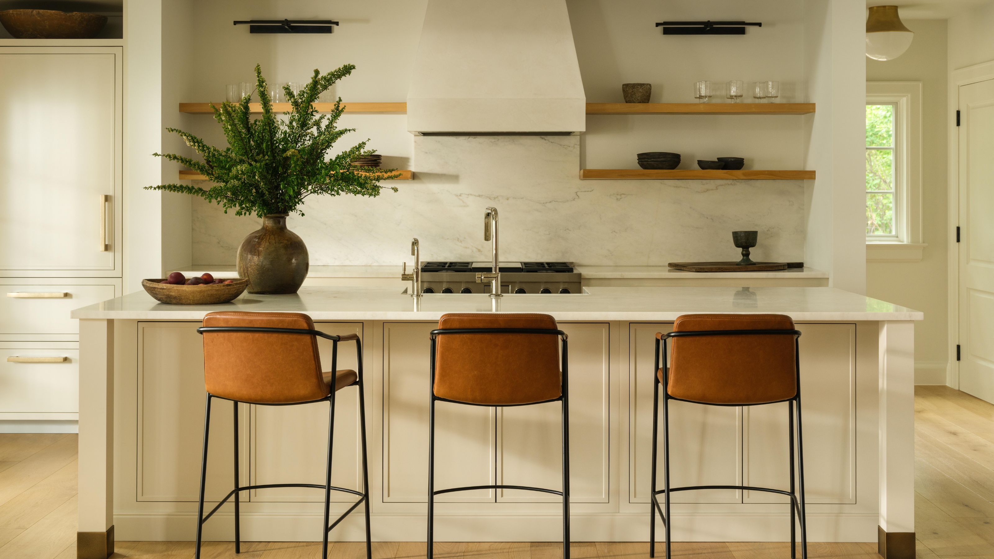 an open plan white shaker kitchen with simple oak shelves, plaster cooker hood, small island and leather and black stools