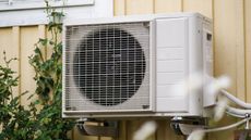 A metal AC Unit mounted to a yellow painted metal house wall 