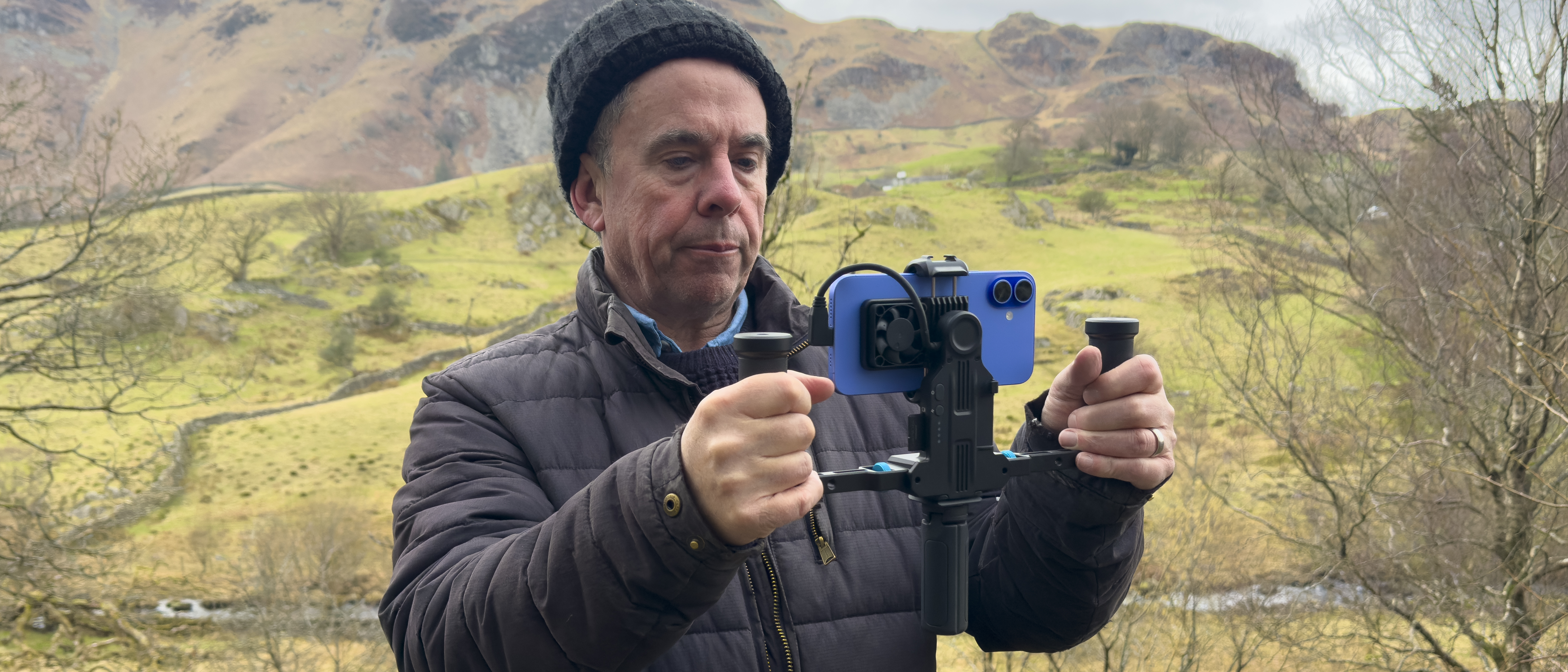 Photographer holding a dual-handle smartphone video rig with mounted phone while filming in a mountainous landscape.