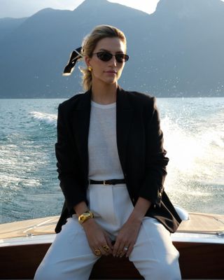 Woman wears black blazer with white T-shirt, white pants, black skinny belt, and black '90s sunglasses while on boat.