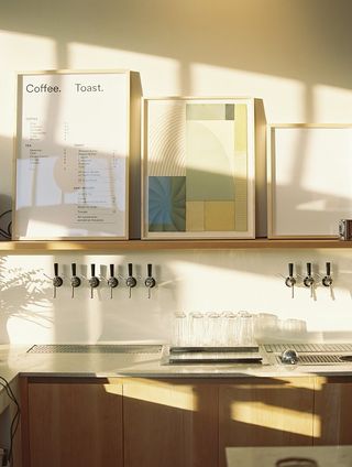 A sun-lit coffee bar counter features wooden fixtures and a series of wall art pieces hanging on its top shelf.