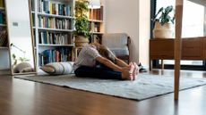 woman sitting on the floor with legs outstretched in a forward fold stretch and head turned down. she's on a large square rug in a home with a book case behind her