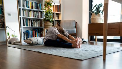 woman sitting on the floor with legs outstretched in a forward fold stretch and head turned down. she's on a large square rug in a home with a book case behind her