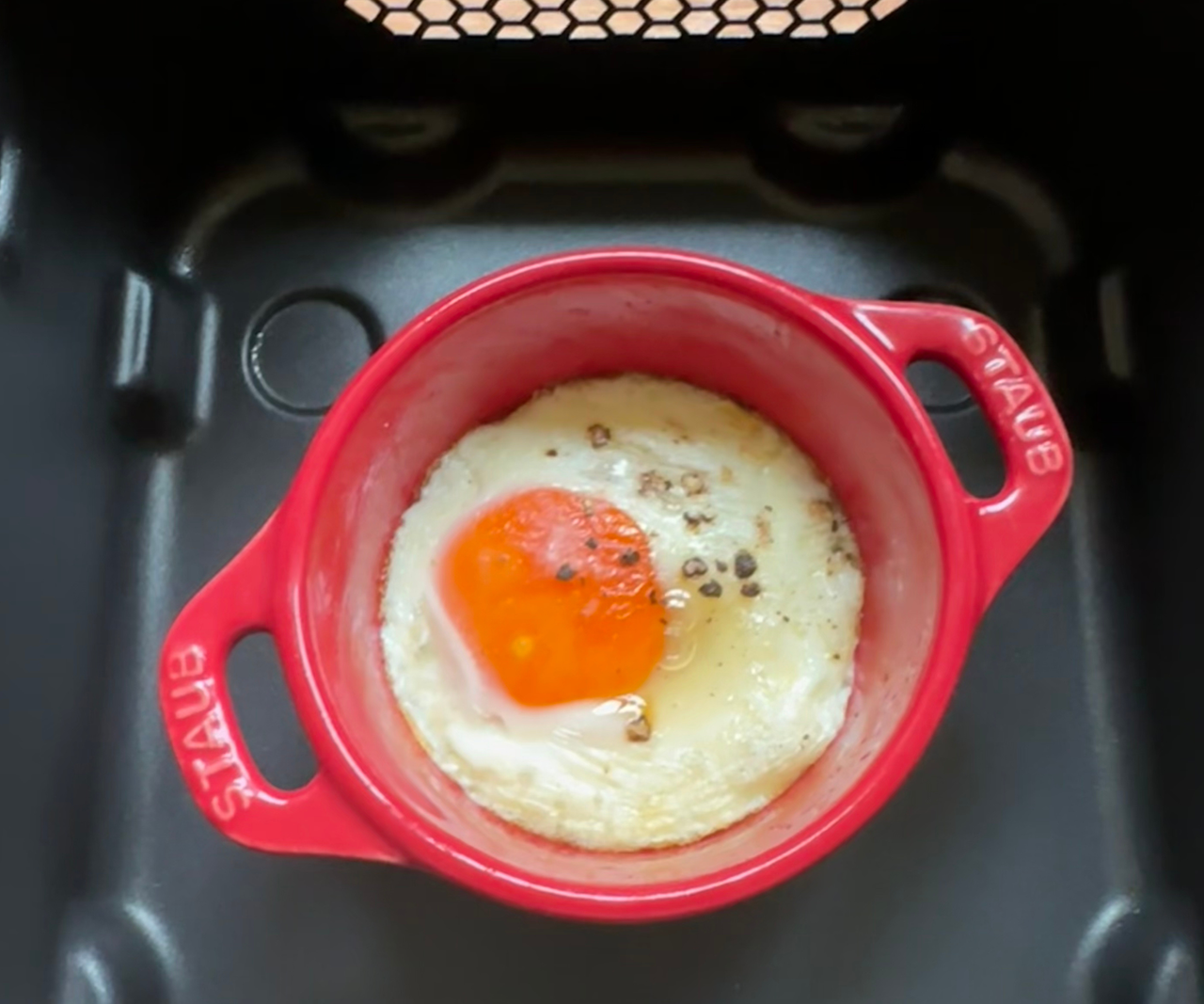 A small red STAUB ramekin inside a black air fryer basket, with a poached egg inside.