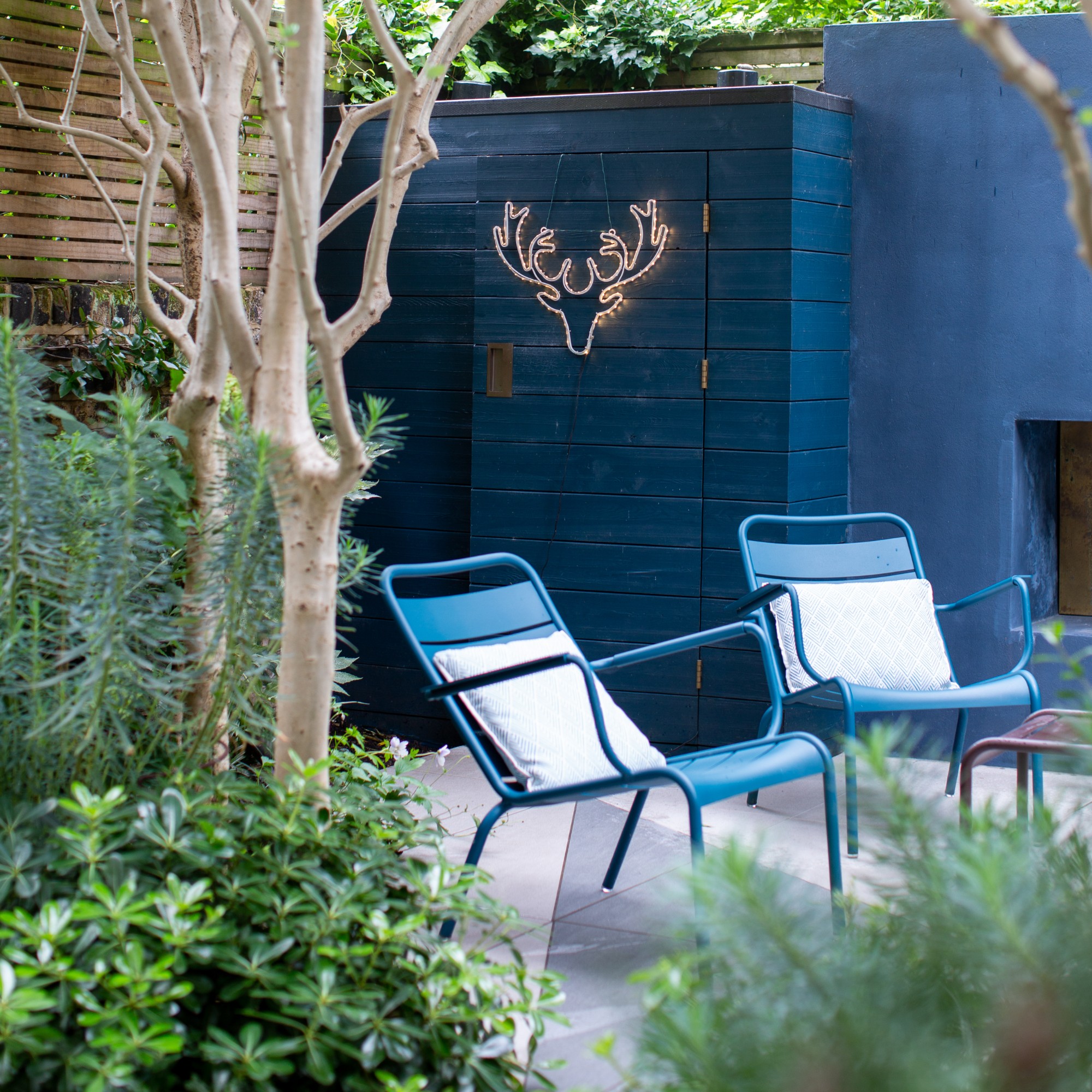 A garden with a blue painted wall and two blue metal garden chairs with greenery and trees around