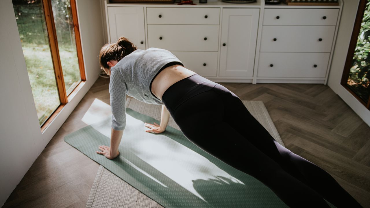 A woman practises yoga / pilates in a sunny room. She holds a difficult plank position, toning the muscles in her abdomen