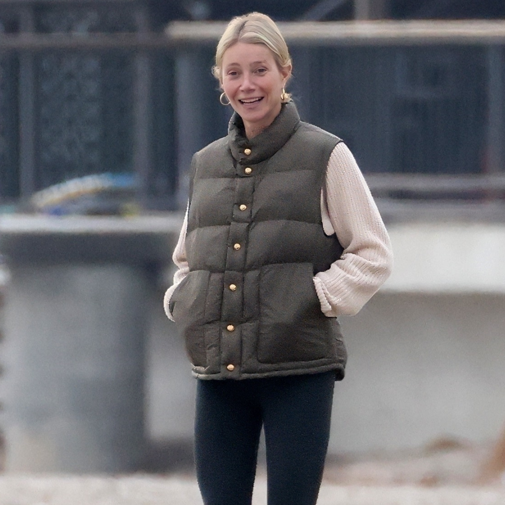 Gwyneth Paltrow walks down the beach wearing black leggings with olive green wellies and an olive green vest top and a beige jumper with gold hoop earrings.