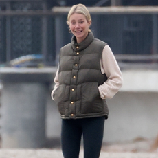 Gwyneth Paltrow walks down the beach wearing black leggings with olive green wellies and an olive green vest top and a beige jumper with gold hoop earrings.