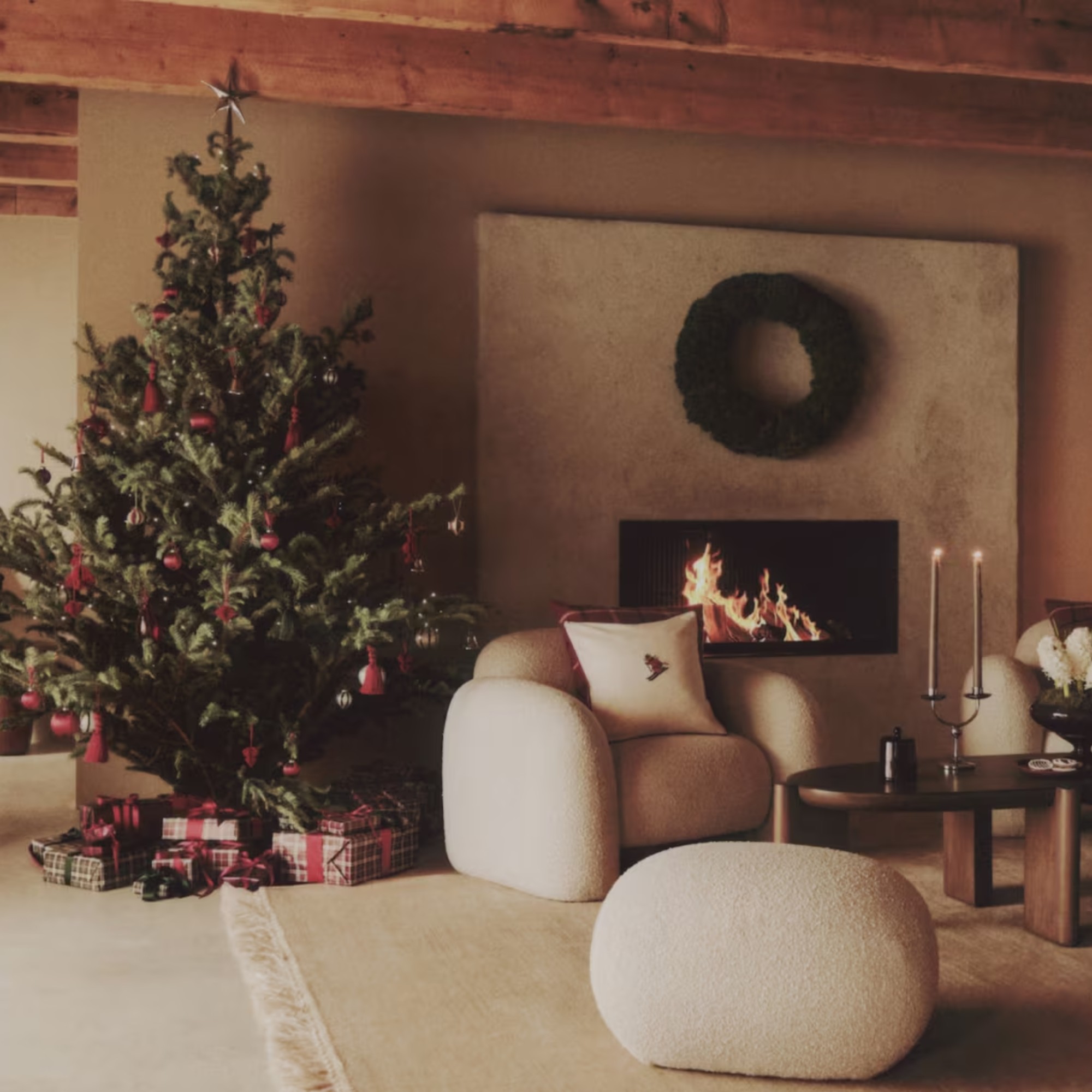 A living room with exposed beams and a lit fireplace with a decorated Christmas tree and a wreath hung above the fireplace