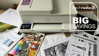 A HP Envy 6555e printer during our review, with freshly printed paper scattered around, and a TechRadar badge that reads 'Big Savings'