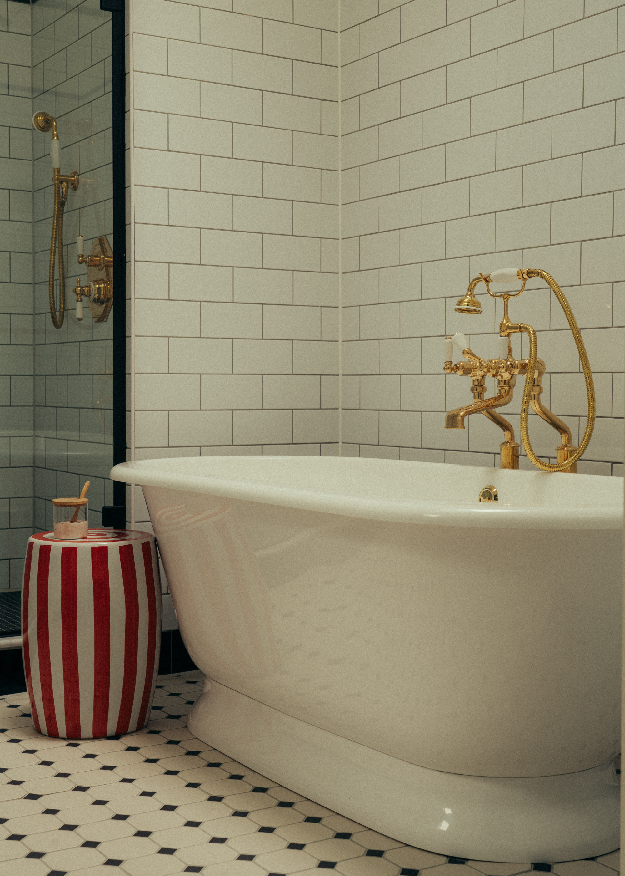 A white tiled bathroom with a large tub, gold hardware, and a red and white striped stool with a bottle of bath salts
