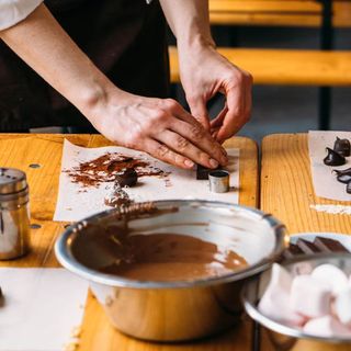 A lifestyle shot of a woman's hands making chocolate behind a large bowl of melted chocolate