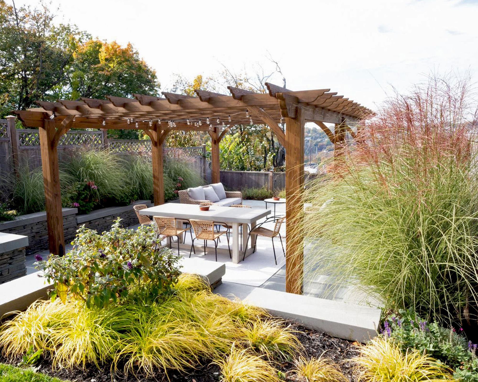 Freestanding wooden pergola on patio with garden furniture surrounded by immersive planting of ornamental grasses and shrubs in raised beds