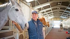 Top showjumper William Funnell stands in an indoor barn in front of a stable with his hand resting underneath a greay horse&rsquo;s head.