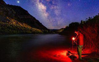 Photographer at Night Capturing Astrophotography Milky Way Landscape - Man with headlamp photographing a bright galaxy along scenic canyon and river with dark skies.