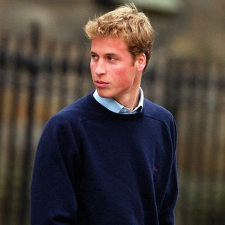 An early 2000s picture of Prince William in a navy sweater looking behind him in front of a fence at St. Andrews