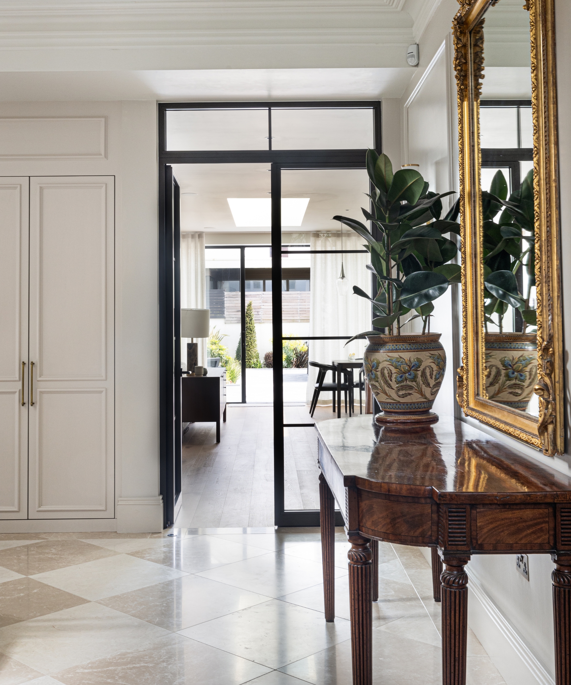 Antique wooden console table with potted plant and gilt mirror in hallway, checkered floor, glazed black doors.