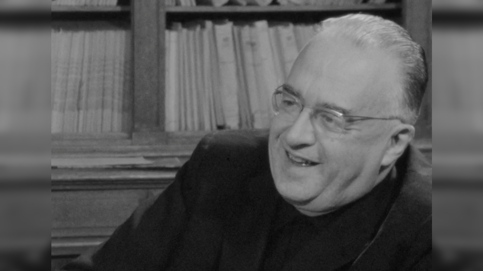 A still of Georges Lemaître smiling and talking and sitting in front of a bookshelf from the rediscovered video.