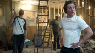 Ebon Moss-Bachrach looking at the ceiling while holding a broom and Jeremy Allen White standing with his hands on his hips. The Beef is torn a part behind them.