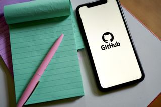 GitHub logo on a smartphone placed down on a desk next to a green notepad and pink pen
