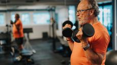 older man facing sideways wearing an orange tshirt in the forefront of the photo performing a biceps curl with two dumbbells in a gym setting. there is another man doing the same, blurred in the background.