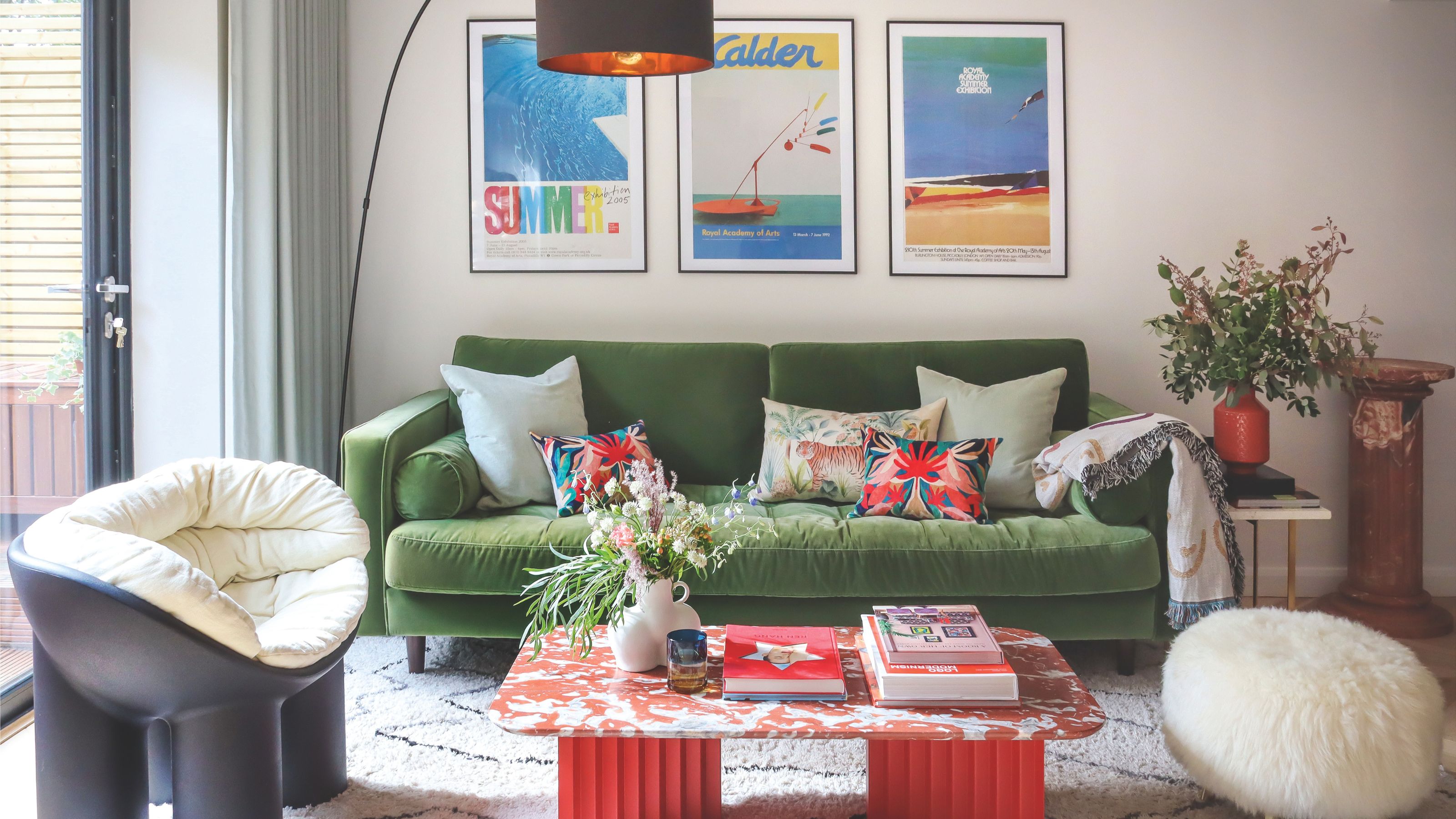 A vibrant, modern living room featuring a green velvet sofa, a red marble-topped coffee table, and three framed art posters on a white wall.