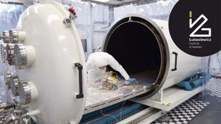 Thermal Vacuum Chamber testing of Structural-Thermal Propulsion Module for NanoSatellites. The chamber looks like a giant white capsule with one end pulled out revealing a table inside. A person in a white, protective bodysuit (covered from head to toe) is inspecting the item inside the chamber and it's many, many wires).