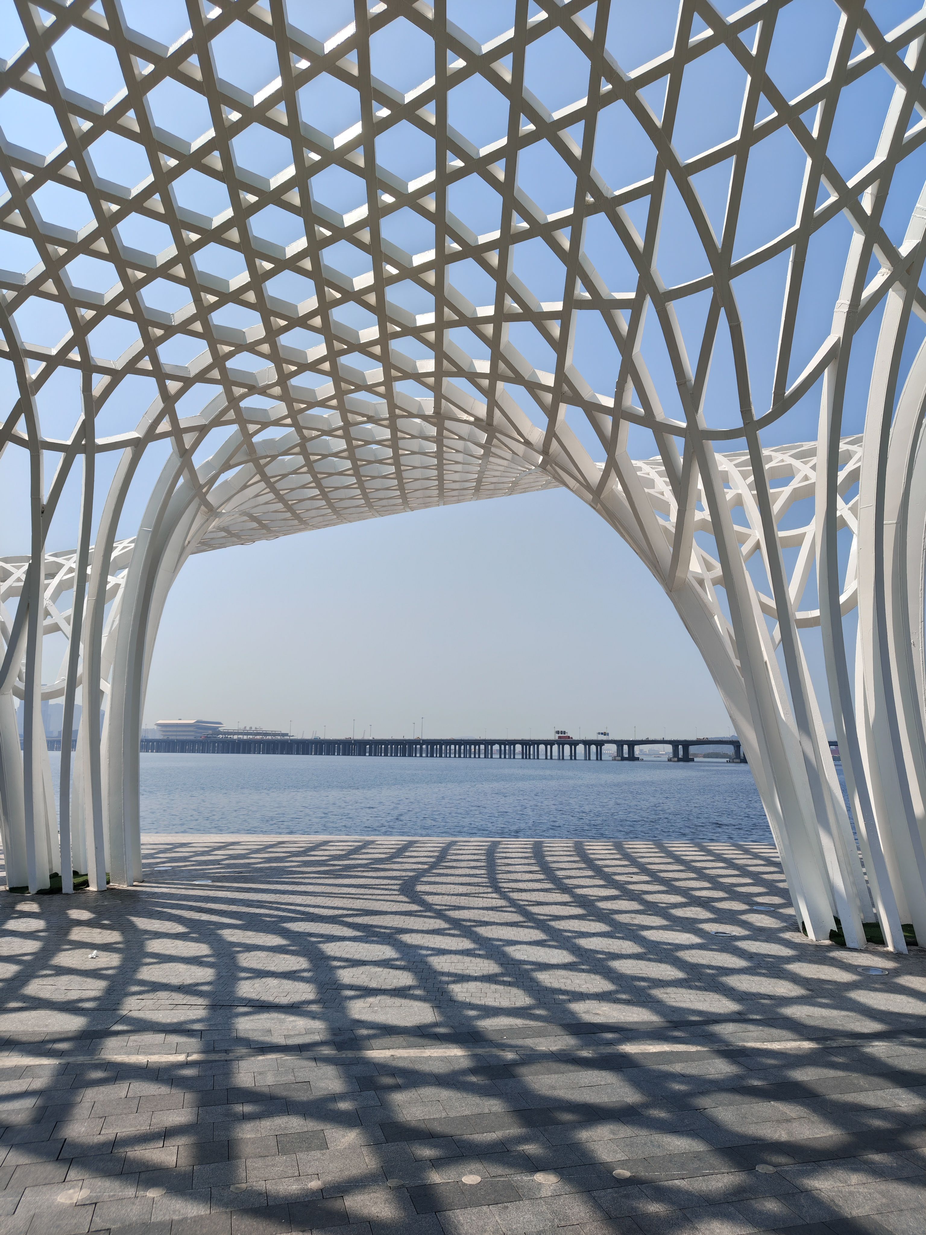 A view through a large, white, lattice-like architectural canopy with a complex geometric diamond pattern. The structure casts a bold, distorted shadow onto the gray stone tiles below. In the distance, a long bridge stretches across a calm blue body of water under a clear, bright sky.