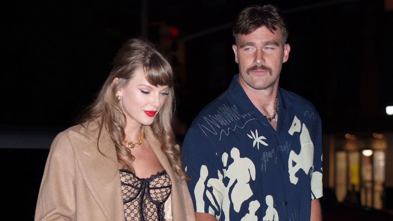 Taylor Swift wearing a tan coat and bustier walking at night next to Travis Kelce who is wearing a blue wildly patterned shirt