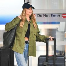 Margot Robbie wears a yankees baseball cap, green shirt, blue straight-leg jeans, black ankle boots, a black shoulder bag and carries away luggage.