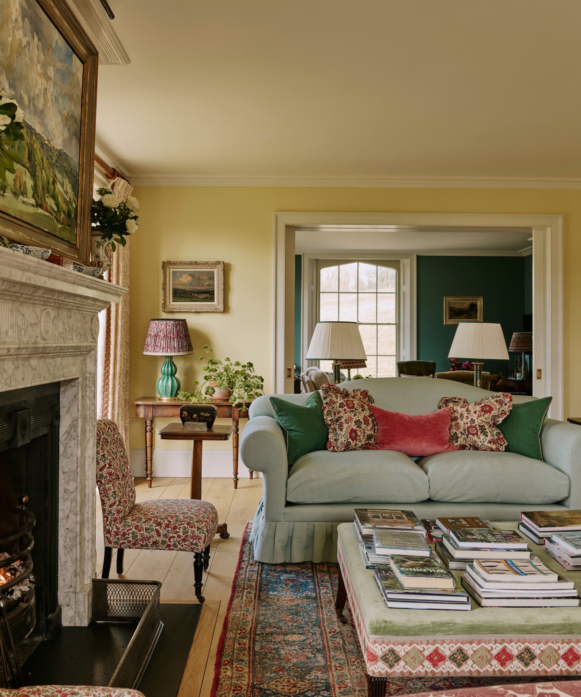 elegantly styled living room featuring a light blue skirted sofa with colorful accent pillows, a marble fireplace, an upholstered coffee table stacked with books, and an doorway opening into a teal room