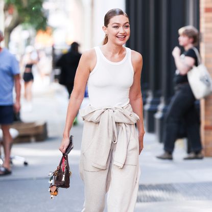a blonde woman in a white tank top smiling at the camera with tan pants and a brown bag