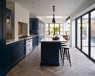 A long kitchen extension with parquet flooring and dark blue cabinets with a central island with bar stools