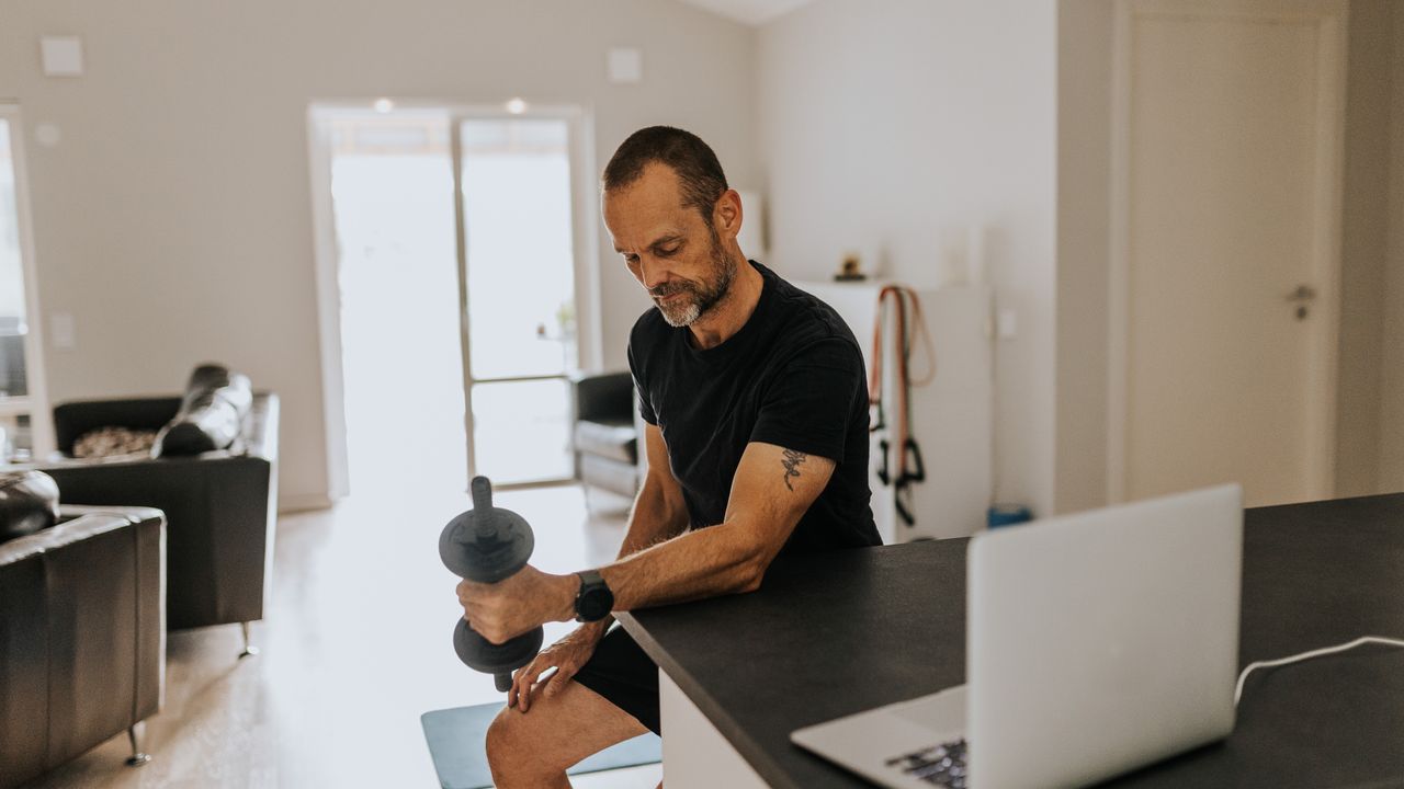 A man in a t-shirt and shorts sits at a breakfast bar in his home doing a bicep curl. His forearm rests on the surface of the bar and his hand grips a dumbbell. Behind him we see two chairs and a large window. There&#039;s also a laptop open on the breakfast bar.