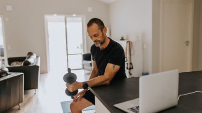 A man in a t-shirt and shorts sits at a breakfast bar in his home doing a bicep curl. His forearm rests on the surface of the bar and his hand grips a dumbbell. Behind him we see two chairs and a large window. There's also a laptop open on the breakfast bar.