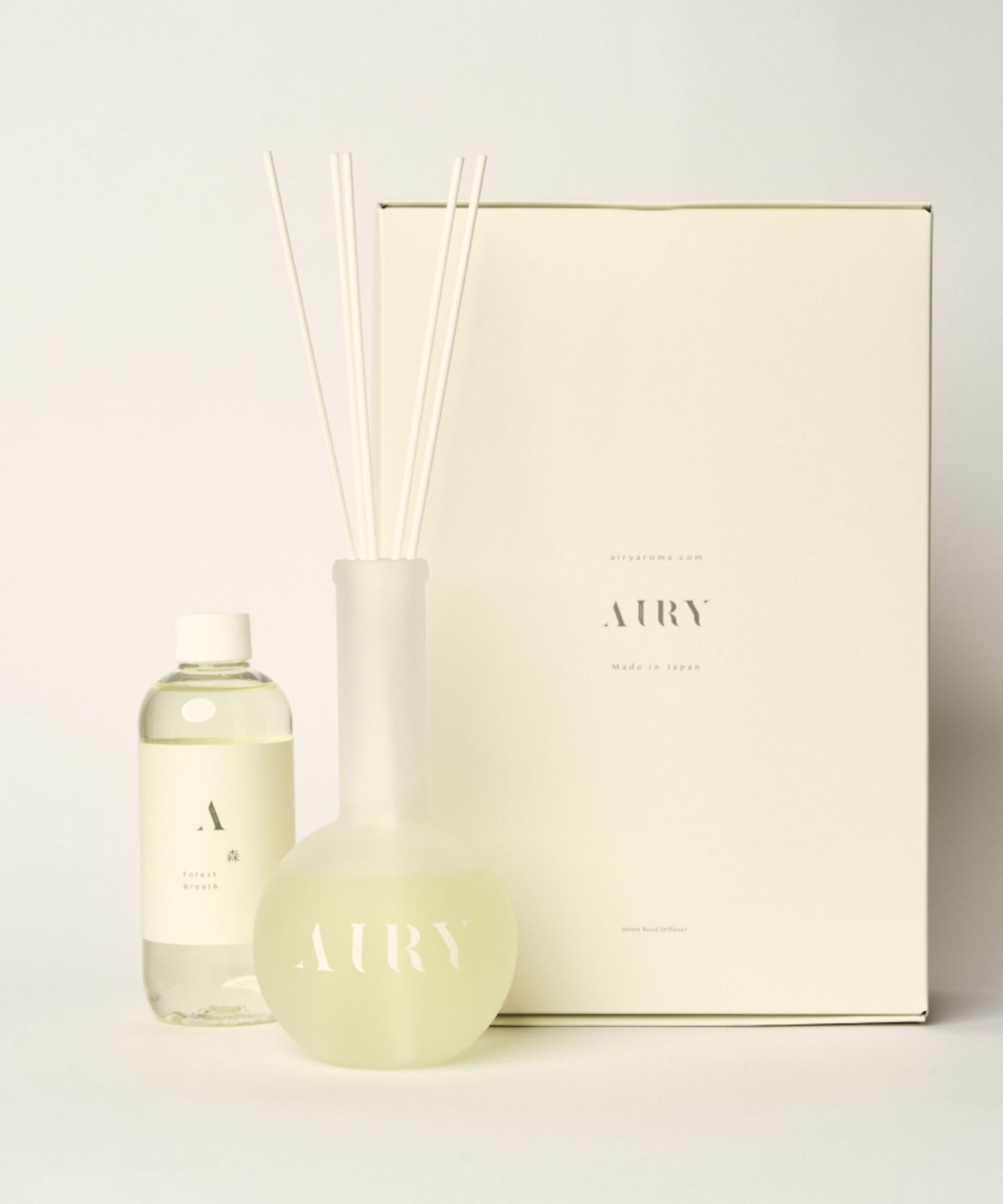 A glass Airy Fragrances reed diffuser filled with white reeds, with a clear bottle of refillable fragrance with a white lid, and a large cardboard packaging box