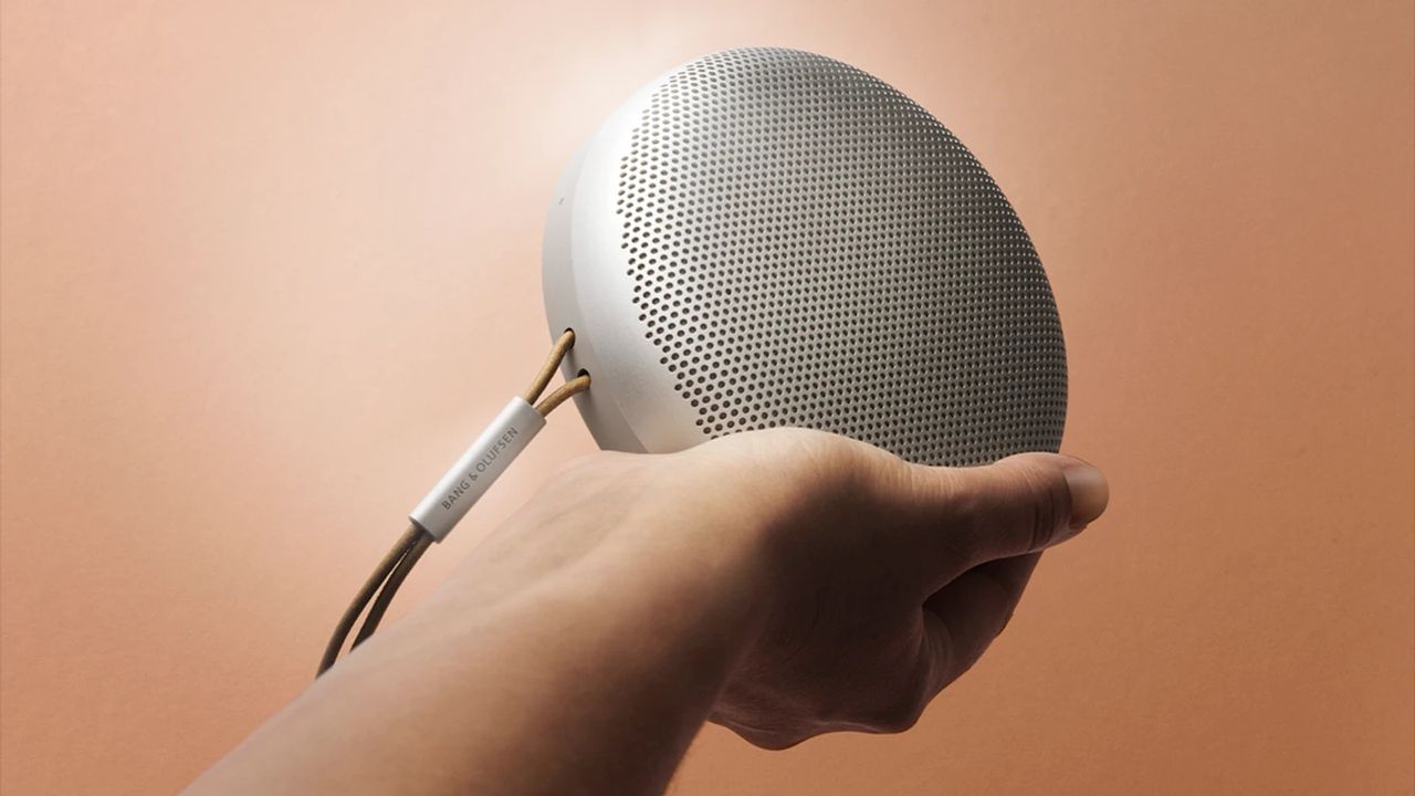 A silver Bang and Olufsen Beosound A1 (2nd gen) speaker being held in an outstretched hand. The background is a peachy beige colour.