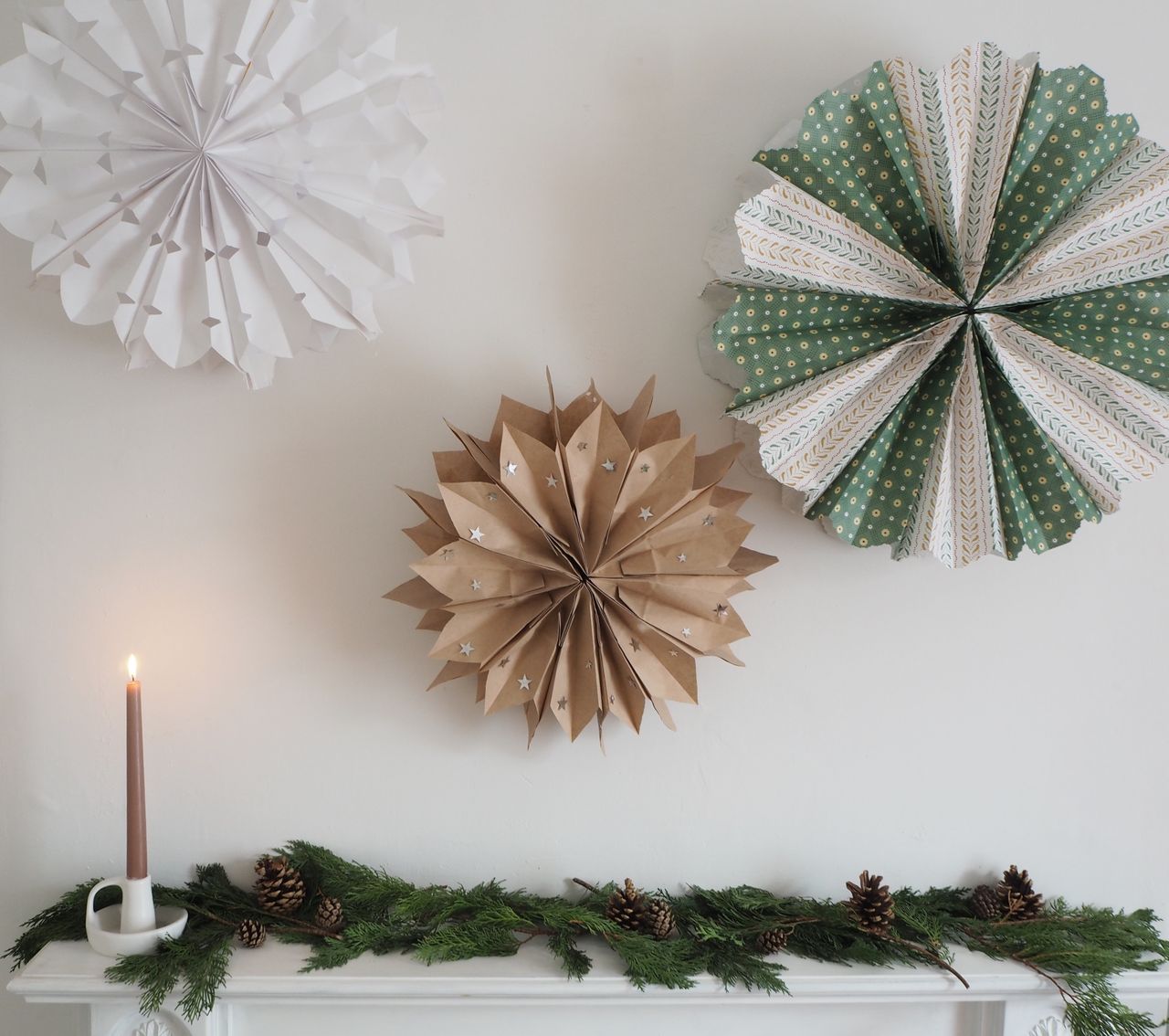 Christmas paper bag decorations on a wall above mantel, foliage on mantel, pine cones, single lit candle 