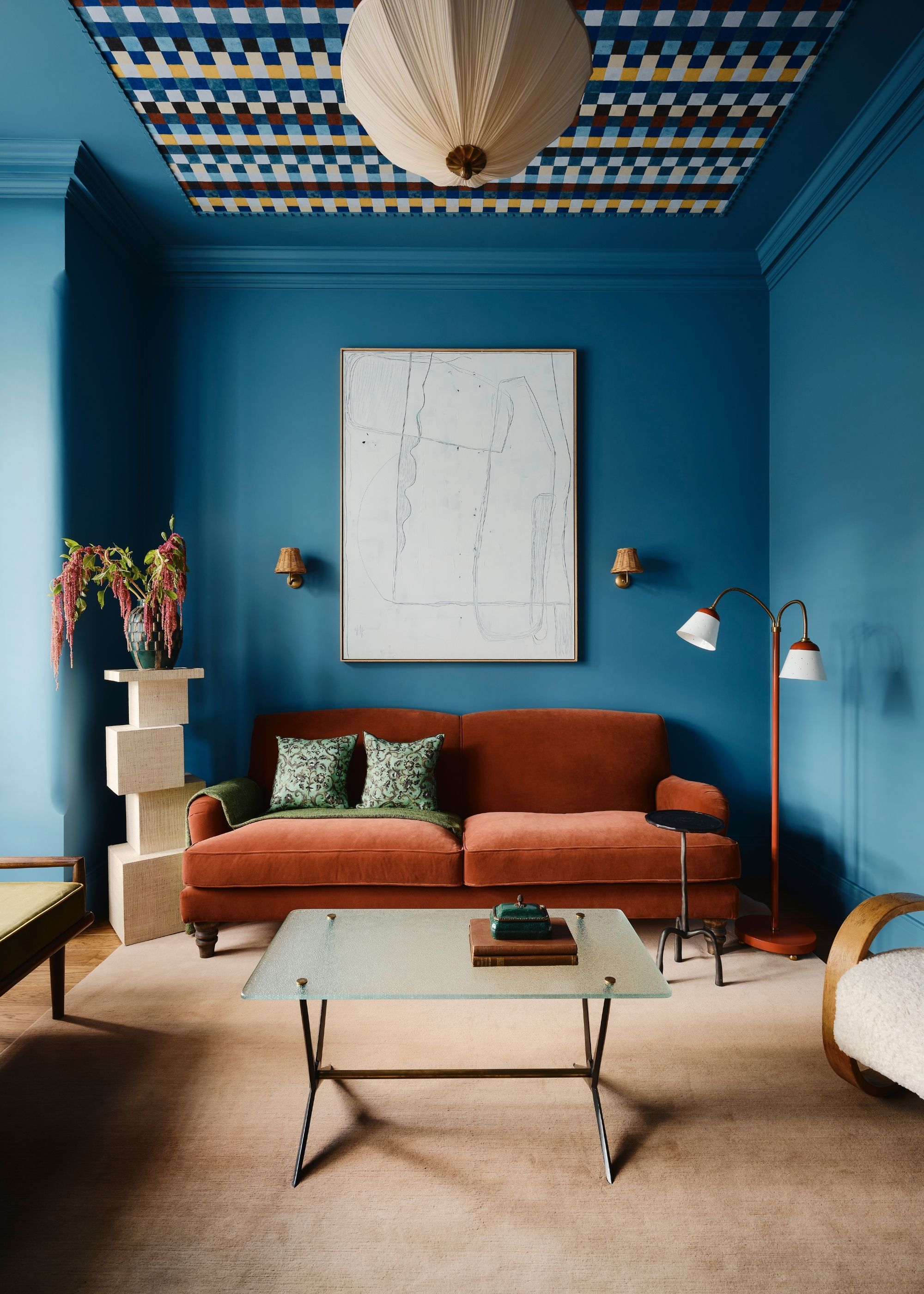 A modern, period living room with mid-blue color-drenched walls and a geometric-patterned wallpaper on the ceiling. A terracotta-colored sofa and a beige carpet with minimal, modern furniture.