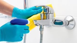 Someone cleaning a bathroom faucet with a spray bottle and cloth while wearing gloves