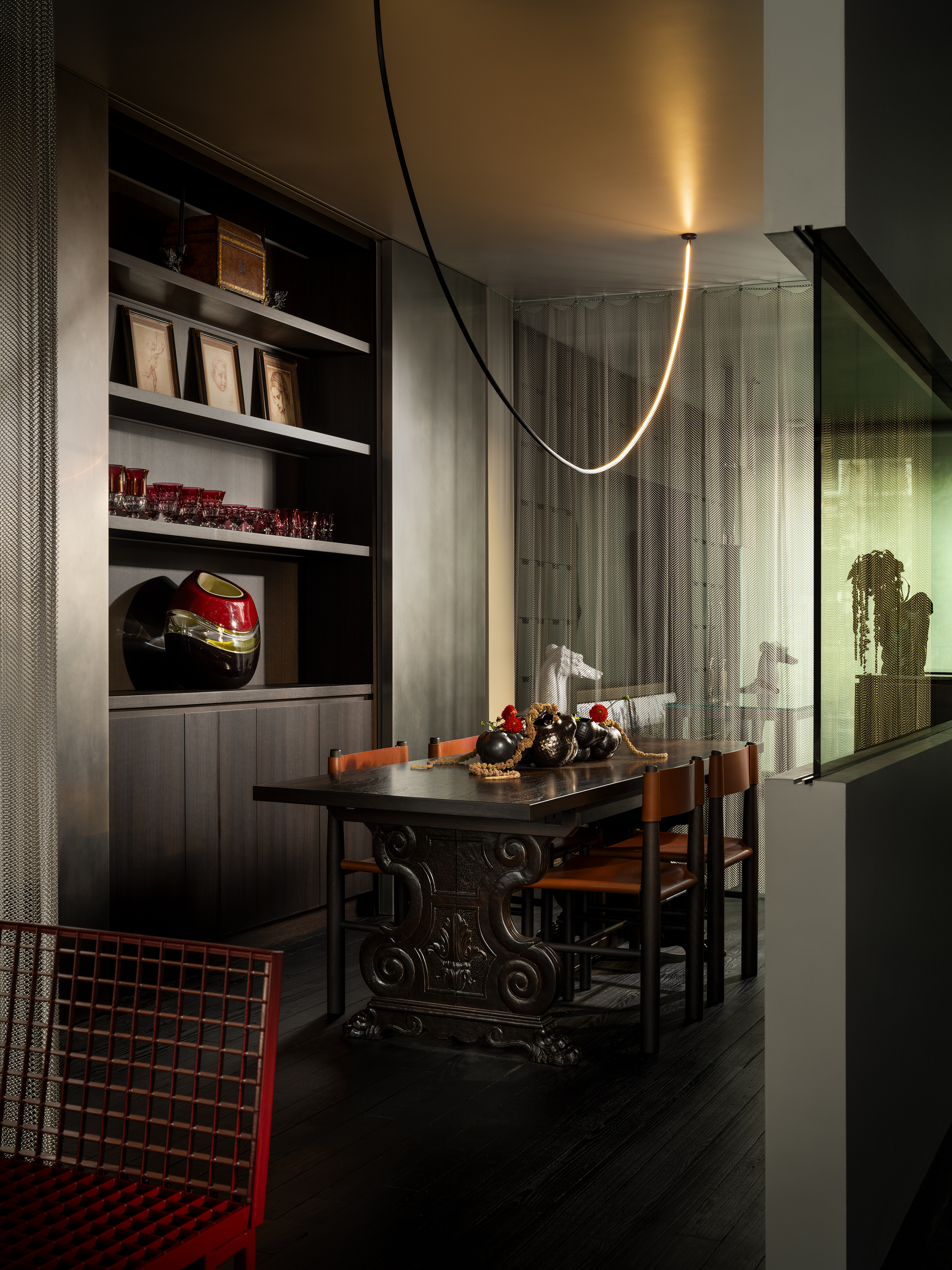 dark and moody dining room with chain mail curtain as room divider, built-in bookshelves in a dark timber styled with picture frames and sculptures, an ornate dark timber dining table with leather chairs, dark floorboards, and a red metal chair in the foreground