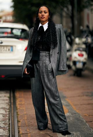 A woman wearing geek chic trend in the form of a grey blazer and grey trousers with a white collar shirt, black vest, and black boots during Milan Fashion Week spring/summer 2025.