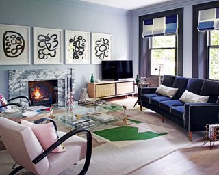 A blue living room with pale blue walls, a marble fireplace, purple window frames, dark blue sofa and pale pink armchairs.