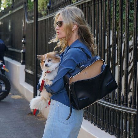 Kate Moss stand outside in London carrying a dog in her arms with a Loewe bag over her shoulder. She wears a light blue denim skirt with black French-style strappy heels.