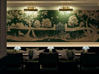 A gold-tinted hotel lounge features bucolic wall murals in green and cream, mushroom-shaped table lamps in white glass, stripy cushions, and velvety green banquettes.